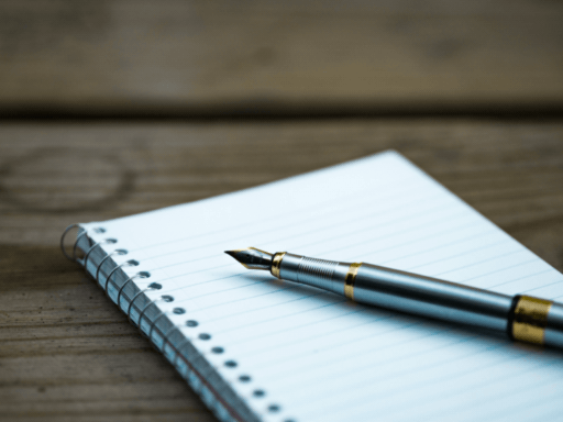 A wooden table with a fountain pen laid on top of a ruled spiral bound notebook